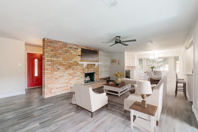 living area with visible vents, a brick fireplace, wood finished floors, baseboards, and ceiling fan with notable chandelier