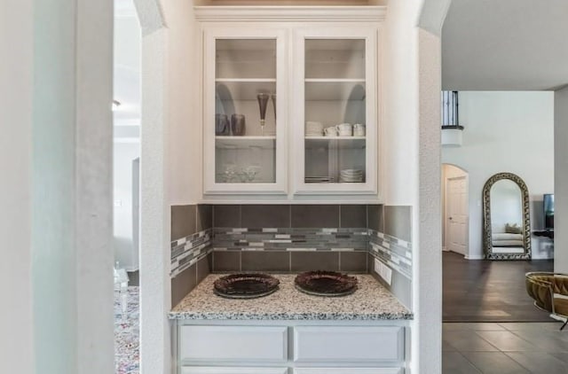 kitchen featuring arched walkways, white cabinets, glass insert cabinets, light stone counters, and backsplash