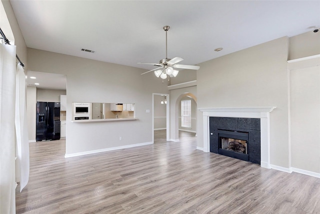 unfurnished living room with a tiled fireplace, arched walkways, light wood finished floors, and ceiling fan