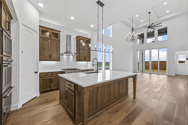 kitchen featuring a center island with sink, hanging light fixtures, light stone countertops, sink, and wall chimney range hood