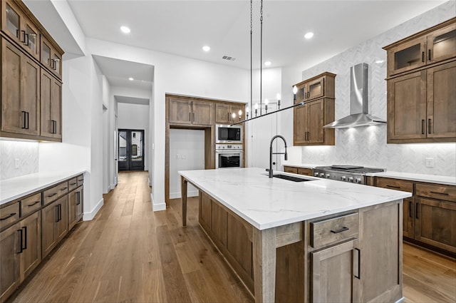 kitchen featuring appliances with stainless steel finishes, hanging light fixtures, an island with sink, light stone counters, and wall chimney range hood