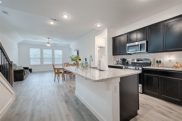 kitchen featuring appliances with stainless steel finishes, decorative backsplash, light hardwood / wood-style floors, light stone countertops, and a center island with sink