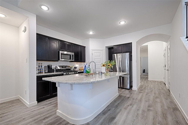 kitchen with sink, light wood-type flooring, appliances with stainless steel finishes, light stone countertops, and a kitchen island with sink