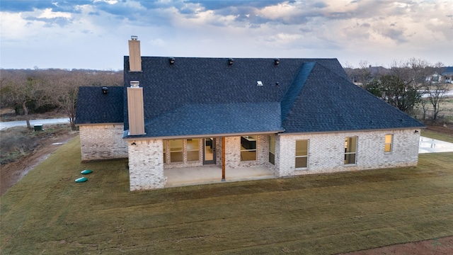 rear view of house with a patio, brick siding, roof with shingles, a lawn, and a chimney