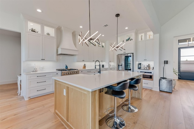 kitchen featuring pendant lighting, appliances with stainless steel finishes, white cabinetry, custom range hood, and a center island with sink