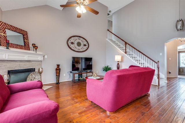 living area featuring a stone fireplace, stairway, wood finished floors, and a ceiling fan