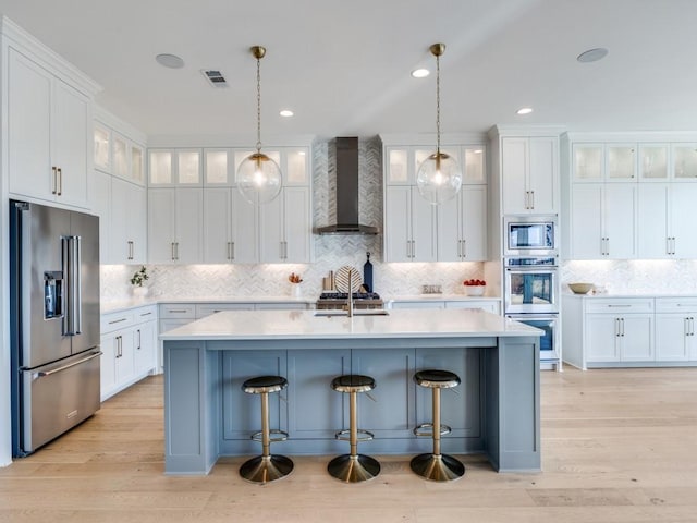 kitchen with white cabinetry, a kitchen breakfast bar, an island with sink, stainless steel appliances, and wall chimney range hood