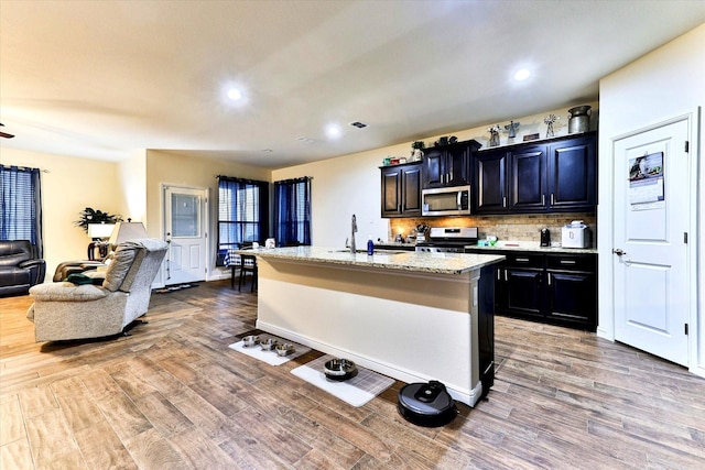 kitchen with sink, appliances with stainless steel finishes, an island with sink, hardwood / wood-style floors, and backsplash