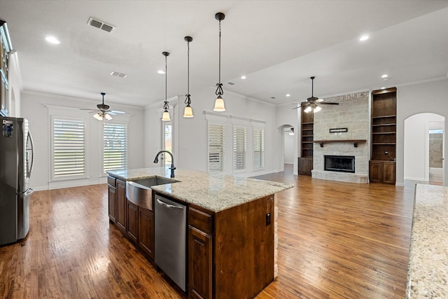 kitchen featuring sink, hanging light fixtures, stainless steel appliances, light stone countertops, and a kitchen island with sink