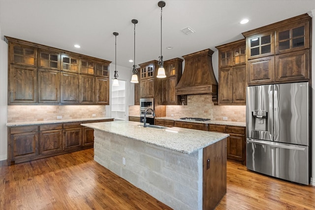 kitchen featuring sink, a kitchen island with sink, stainless steel appliances, light stone counters, and custom range hood