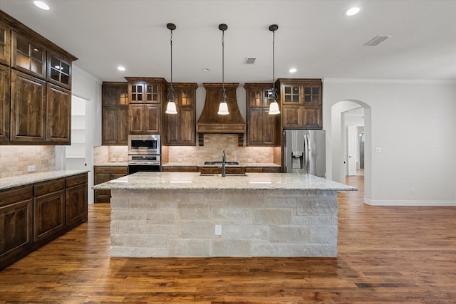 kitchen featuring light stone counters, appliances with stainless steel finishes, a kitchen island with sink, and custom range hood