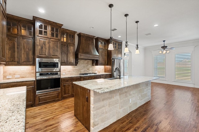 kitchen with an island with sink, custom range hood, pendant lighting, stainless steel appliances, and light stone countertops