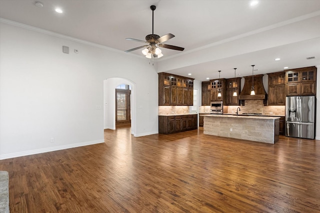 kitchen featuring a center island with sink, appliances with stainless steel finishes, custom range hood, pendant lighting, and backsplash