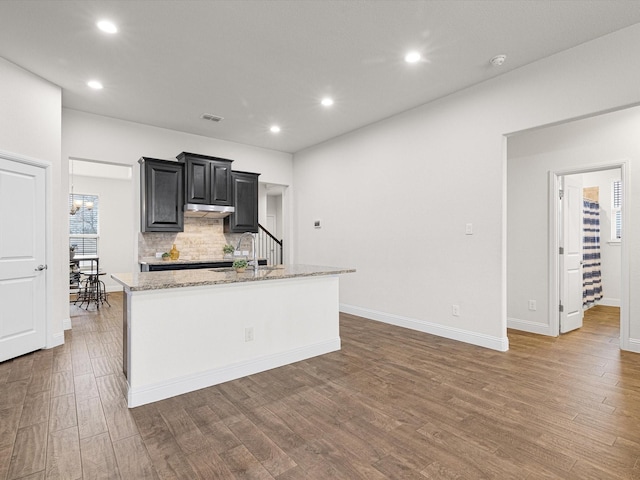 kitchen featuring decorative backsplash, a kitchen island with sink, light stone countertops, dark wood-type flooring, and an inviting chandelier