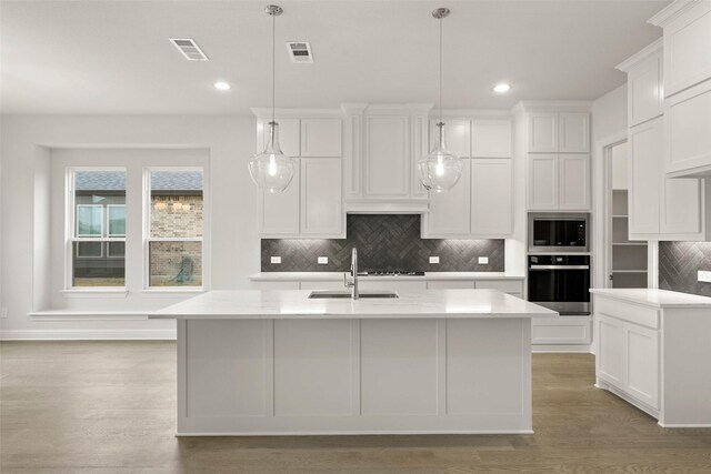 kitchen with stainless steel appliances, white cabinetry, a center island with sink, light stone countertops, and decorative light fixtures