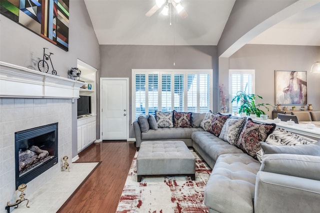 living room featuring built in shelves, high vaulted ceiling, hardwood / wood-style flooring, ceiling fan, and a tiled fireplace