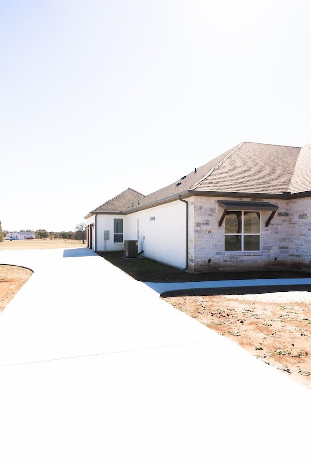 view of front facade with stone siding, roof with shingles, and central air condition unit