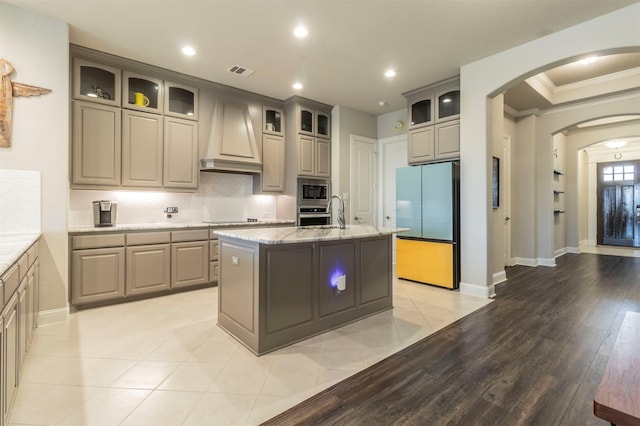 kitchen with appliances with stainless steel finishes, backsplash, a kitchen island with sink, light stone countertops, and custom range hood