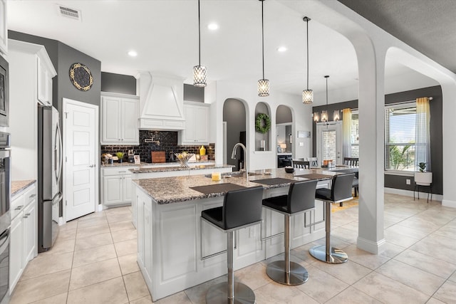 kitchen with a center island with sink, custom exhaust hood, stainless steel appliances, visible vents, and light stone countertops