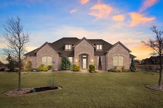 french country inspired facade featuring roof with shingles, brick siding, and a front lawn