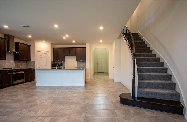 kitchen featuring tasteful backsplash, an island with sink, stainless steel oven, dark brown cabinets, and wall chimney exhaust hood