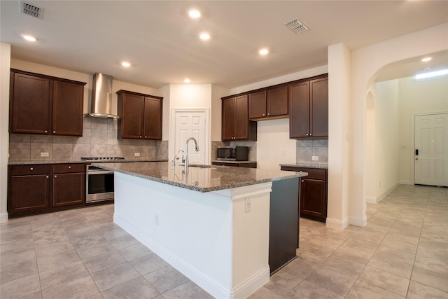 kitchen featuring sink, dark stone countertops, oven, a kitchen island with sink, and wall chimney range hood