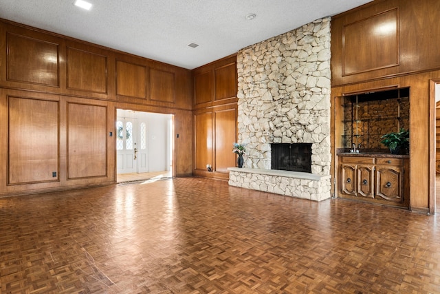 unfurnished living room with wooden walls, dark parquet flooring, a stone fireplace, and a textured ceiling