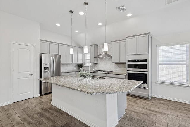 kitchen featuring appliances with stainless steel finishes, decorative light fixtures, a kitchen island with sink, light stone counters, and wall chimney range hood