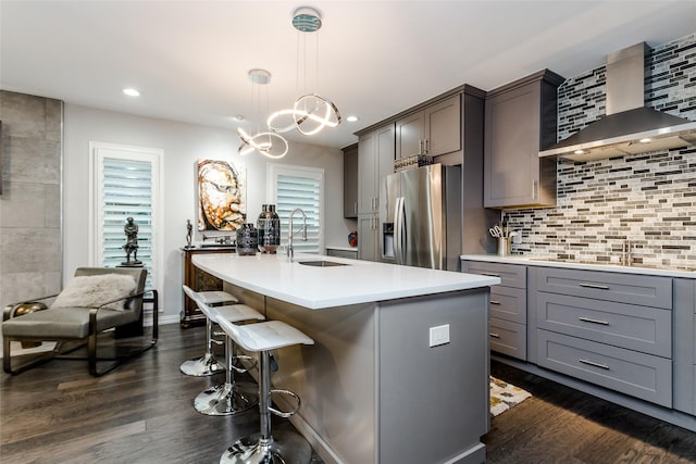 kitchen featuring sink, stainless steel fridge with ice dispenser, a center island with sink, pendant lighting, and wall chimney range hood