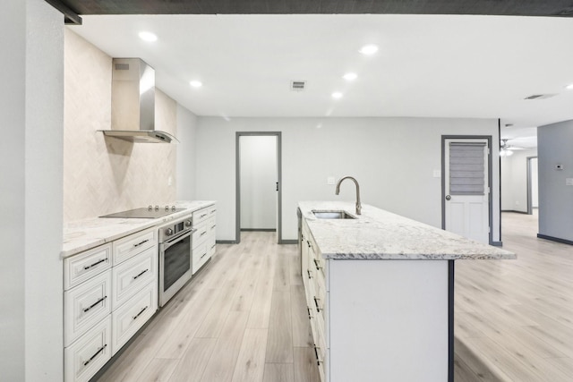 kitchen with sink, a center island with sink, black electric cooktop, oven, and wall chimney exhaust hood