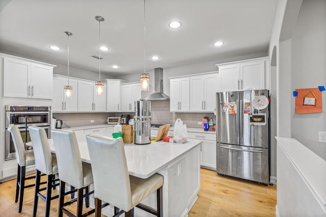 kitchen featuring hanging light fixtures, wall chimney range hood, a kitchen island, stainless steel appliances, and white cabinets