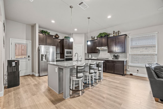 kitchen with a kitchen island with sink, stainless steel appliances, dark brown cabinetry, decorative light fixtures, and decorative backsplash