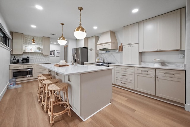 kitchen with decorative light fixtures, appliances with stainless steel finishes, custom range hood, a kitchen island with sink, and white cabinets