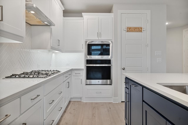 kitchen with wall chimney exhaust hood, light hardwood / wood-style flooring, appliances with stainless steel finishes, white cabinets, and backsplash