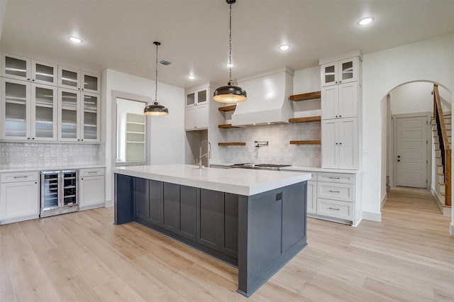 kitchen with sink, premium range hood, a kitchen island with sink, white cabinets, and beverage cooler