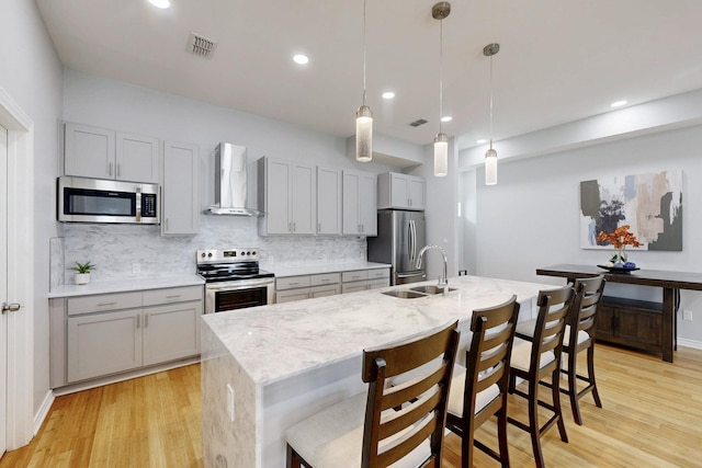 kitchen with hanging light fixtures, appliances with stainless steel finishes, light stone countertops, wall chimney exhaust hood, and a center island with sink