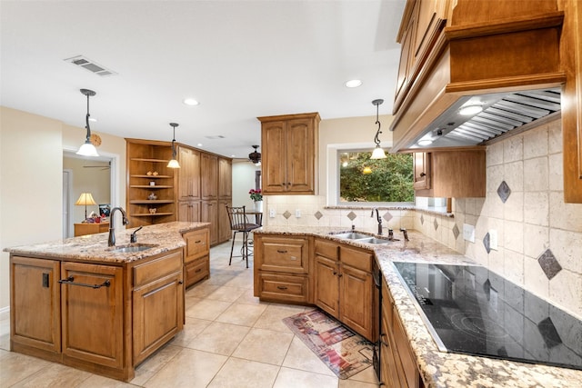 kitchen featuring decorative light fixtures, sink, black electric stovetop, light stone countertops, and a center island with sink