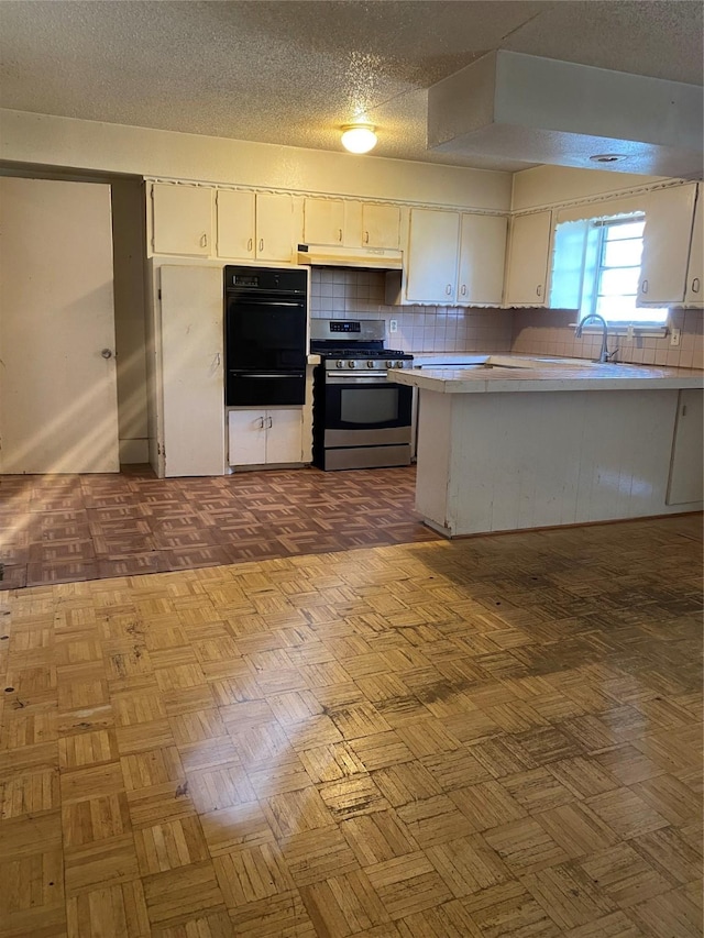 kitchen with tile countertops, stainless steel range with gas stovetop, black oven, white cabinets, and backsplash