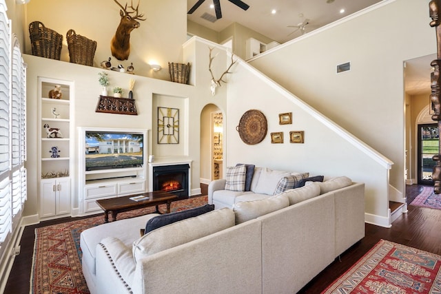 living room with ceiling fan, a towering ceiling, built in features, and dark hardwood / wood-style flooring