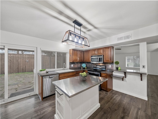 kitchen featuring stainless steel appliances, a sink, brown cabinetry, dark countertops, and decorative light fixtures