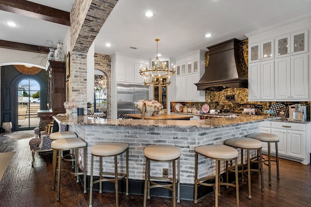 kitchen with premium range hood, built in refrigerator, decorative light fixtures, light stone countertops, and white cabinets