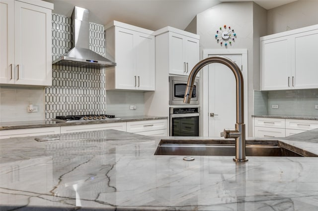 kitchen with light stone countertops, appliances with stainless steel finishes, wall chimney range hood, and white cabinets