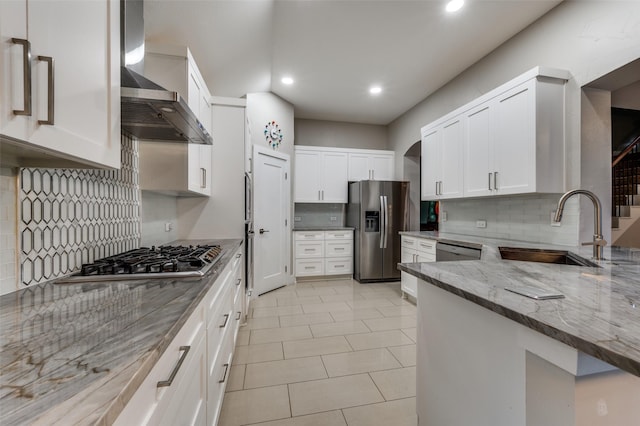 kitchen with white cabinets, stainless steel appliances, light stone counters, wall chimney exhaust hood, and sink