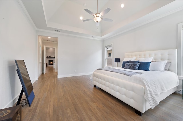 bedroom with a warm lit fireplace, wood finished floors, baseboards, a raised ceiling, and crown molding