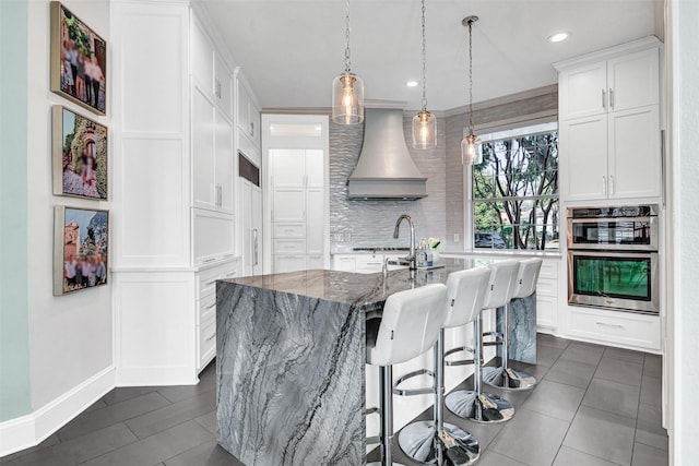 kitchen featuring stone counters, white cabinetry, tasteful backsplash, custom range hood, and a center island with sink