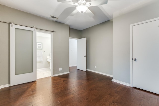 unfurnished bedroom with a barn door, dark hardwood / wood-style floors, connected bathroom, and ceiling fan