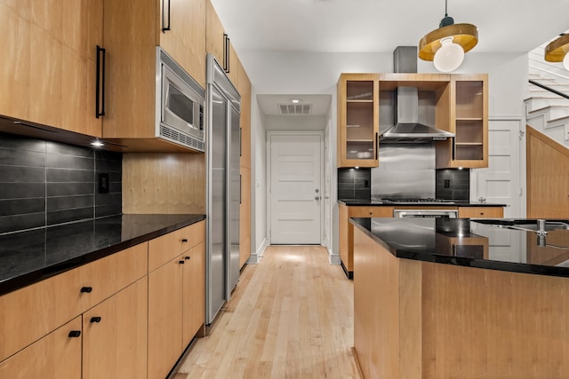 kitchen with wall chimney exhaust hood, light hardwood / wood-style flooring, pendant lighting, stainless steel appliances, and backsplash