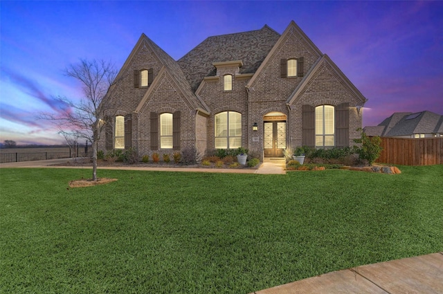 french country inspired facade featuring a shingled roof, brick siding, fence, and a front lawn