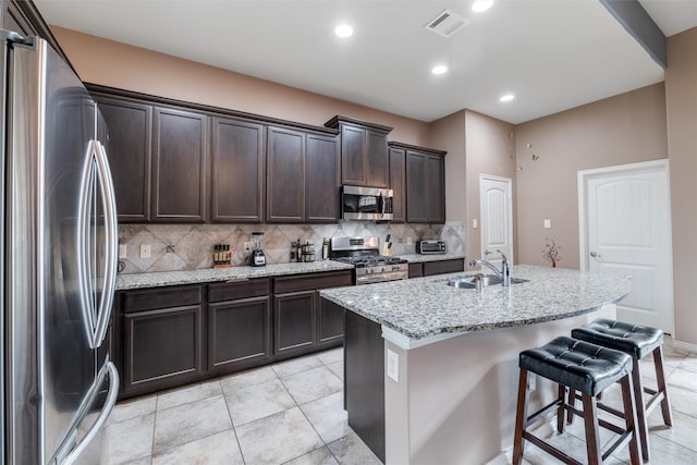 kitchen featuring tasteful backsplash, an island with sink, dark brown cabinetry, stainless steel appliances, and light stone countertops