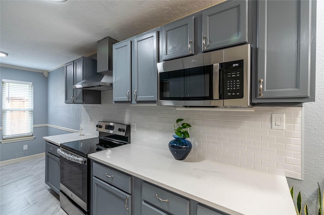 kitchen featuring gray cabinets, appliances with stainless steel finishes, decorative backsplash, wall chimney range hood, and a textured ceiling